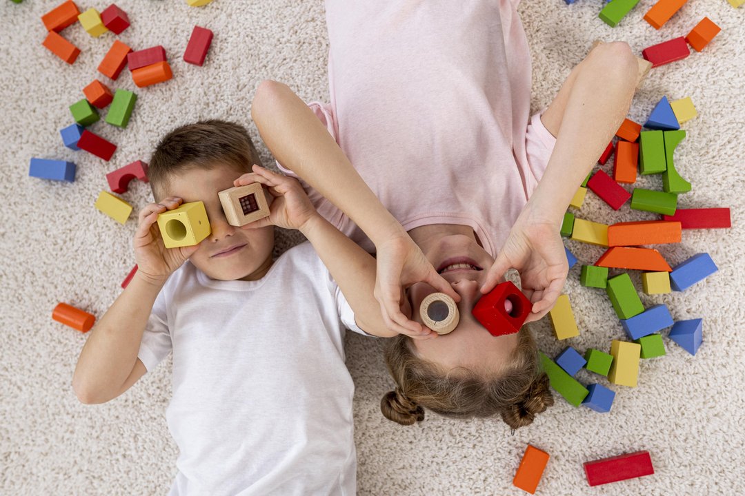 Zwei Kinder liegen am Boden und blicken durch Bausteine, die sie wie Brillen ansetzen