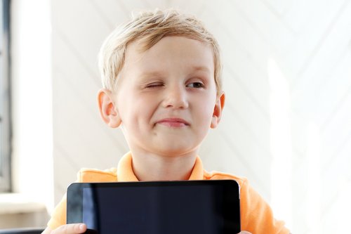 Ein Junge blickt schmunzelnd zur Seite und kneift ein Auge zu, während er ein Tablet in der Hand hält