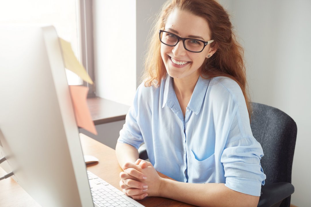 Eine junge Frau sitzt vor dem PC-Bildschirm und lächelt erleichtert in die Kamera