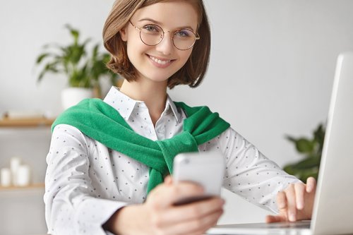 Eine Kita-Leiterin sitzt lächelnd am PC, mit Blick in die Kamera, das Smartphone in der Hand